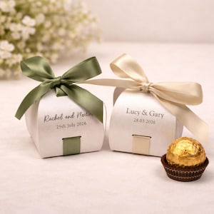 Peut inclure: Deux boîtes à cadeaux blanches avec des nœuds en satin, une verte olive et une crème. Chaque boîte porte un nom et une date imprimés. Un seul bonbon au chocolat dans une coupelle en papier marron est posé à côté d'une boîte.