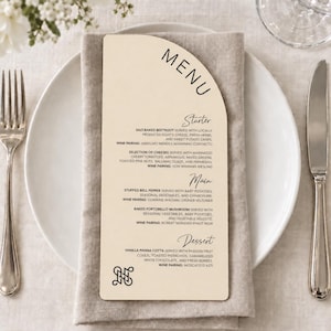 May include: An arched, cream-coloured paper menu card with black text listing courses including salt-baked beetroot, selection of cheeses, stuffed bell pepper, baked portobello mushroom, and vanilla panna cotta, featuring a decorative logo at the bottom.