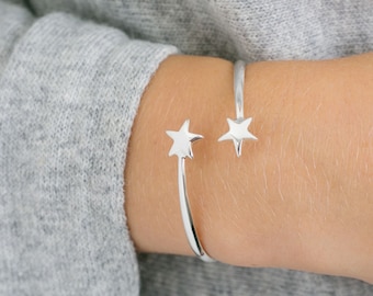 Può includere: Un bracciale aperto color argento con due ciondoli lucidi a forma di stella alle estremità. Il bracciale presenta una finitura metallica liscia e riflettente, progettato per essere indossato attorno al polso con un aspetto semplice.