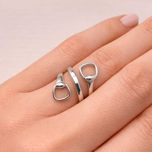 Könnte beinhalten: Ein silberfarbener Ring mit einem einzigartigen Design, das drei offene, abgerundete Formen aufweist. Der Ring umschließt den Finger und erzeugt eine moderne und minimalistische Ästhetik. Der Ring wird auf einer Hand mit hellrosa Nagellack gezeigt.