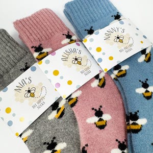 Può includere: Quattro paia di calzini accoglienti in grigio, rosa e blu, ognuno adornato con un motivo ripetuto di bombi. I calzini sono confezionati con etichette che recitano "Bee Happy Socks" e "Bee Cosy Socks".