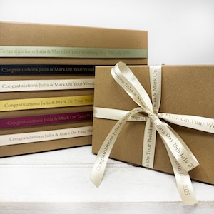May include: A stack of brown gift boxes with beige ribbon tied around the top box. The ribbon has the text "Congratulations Julia & Mark On Your Wedding Day 25th July 2025" printed on it.