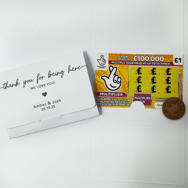 K&ouml;nnte beinhalten: Eine wei&szlig;e Karte mit dem Text "thank you for being here" und "WE LOVE YOU!" liegt neben einem Lotterielos und einem Penny. Das Lotterielos hat eine Multiplikatorfunktion und einen Hauptgewinn von 100.000 &pound;.