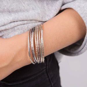 May include: A set of silver, gold, and rose gold bangles with a textured finish. The bangles are stacked on a person's wrist.