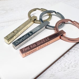 May include: Three gold, silver, and rose gold bar keychains with engraved dates and messages. The keychains are on a white surface.