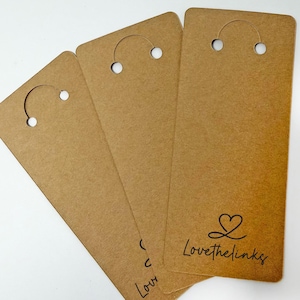 Op de afbeelding: Een stapel bruine rechthoekige hangtags met een gebogen bovenkant en een gat om op te hangen. De tags hebben de tekst "Lovethelinks" met een hartsymbool in zwart gedrukt. De tags zijn gemaakt van dik, getextureerd papier.