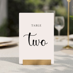 Puede incluir: Tarjeta de número de mesa blanca con caligrafía negra que dice "TABLE two" en un soporte de madera.