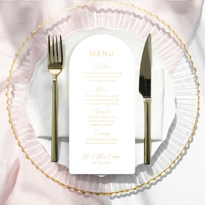 May include: A white card with a gold border and gold text on a white plate with gold trim. The card has a menu for a wedding reception with the following items: Starter, Roasted tomato & sweet red pepper soup with baked sour dough croutons, Main, Beef wellington or wild mushroom ravioli, served with seasonal vegetables, Dessert, Dark chocolate torte served with a sharp raspberry sorbet, Evening, Buffet served from 8pm, Bacon rolls from 11pm, Mr & Mrs Cooper, 26th July 2024.
