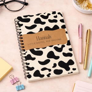 Puede incluir: Un cuaderno con espiral con una cubierta color crema con manchas negras de vaca. Una etiqueta marrón en la parte delantera dice "Hannah" y "Little Notes. Big Dreams." Bolígrafos rosas y dorados, junto con otros artículos de papelería, están colocados alrededor del cuaderno.