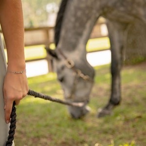 Puede incluir: Una pulsera plateada con dise&ntilde;o de bocado de caballo se lleva en la mu&ntilde;eca. La persona sostiene una cuerda negra, y un caballo gris est&aacute; pastando en el fondo. La escena tiene lugar al aire libre, con una valla de madera y hierba verde.