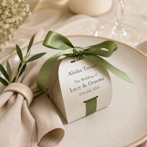 Puede incluir: Una caja de recuerdo color crema atada con una cinta de satén verde salvia. La caja presenta texto negro impreso que dice: Alisha Turner, The Wedding of Lucy & Graeme, 27th July 2026. La caja está colocada sobre un plato de cerámica junto a una servilleta.