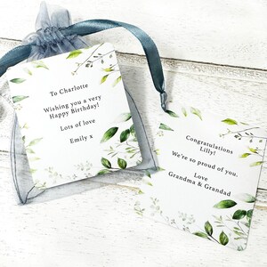 Puede incluir: Dos etiquetas de regalo blancas con detalles de hojas verdes y texto, una en una bolsa azul transparente. Las etiquetas dicen "Para Charlotte, &iexcl;Dese&aacute;ndote un muy feliz cumplea&ntilde;os! Mucho amor, Emily x" y "&iexcl;Felicidades Lilly! Estamos muy orgullosos de ti. Amor Abuela y Abuelo."