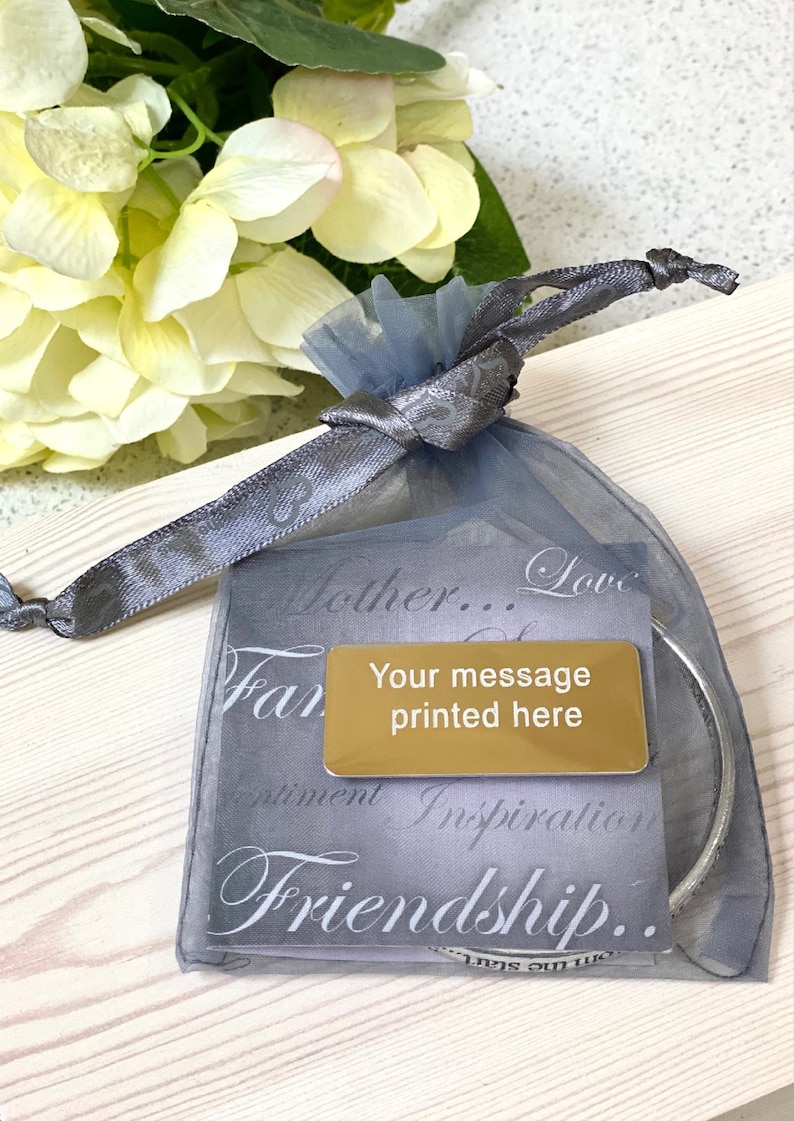 以下が含まれることがあります： グレーのオーガンザバッグに、ゴールドの金属タグが付いています。タグには「メッセージを印刷」と書いてあります。バッグはグレーのリボンで結ばれており、「母...愛...家族...感情...インスピレーション...友情...」という文字が印刷されています。