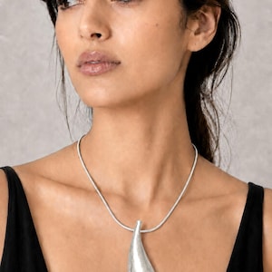 May include: A silver-toned necklace featuring a smooth, elongated teardrop-shaped pendant with a brushed metallic finish. The pendant hangs from a simple, thin, rounded silver-toned chain that rests around the neck, creating a clean and balanced look.