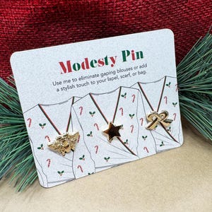 May include: A card with the text "Modesty Pin" and three gold-colored pins: a bee, a star, and a bow. The card also has text that reads "Use me to eliminate gaping blouses or add a stylish touch to your lapel, scarf, or bag."