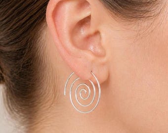 Könnte beinhalten: Ein silberfarbener Ohrring mit einem durchgehenden Spiraldesign. Der Metalldraht bildet eine kreisförmige Spule, die sich zur Mitte hin verjüngt und ein geschichtetes, offenes Erscheinungsbild erzeugt. Der Ohrring ist im Ohrläppchen platziert.