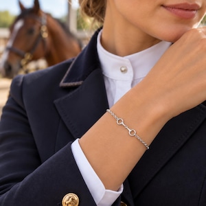 Könnte beinhalten: Ein silberfarbenes Kettenarmband mit einem zentralen Trensen-Glied-Design. Das Armband besteht aus feinen, runden Gliedern und einem sicheren Verschluss und weist eine polierte metallische Oberfläche am Handgelenk auf.