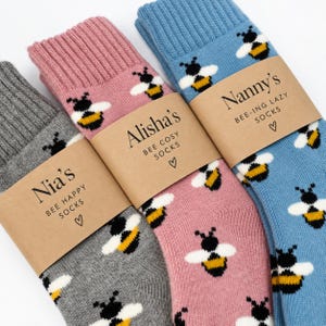 May include: Three pairs of crew socks in gray, pink, and blue, each featuring a repeating bee pattern. Each pair is wrapped in a tan paper band with printed text: Nia's Bee Happy Socks, Alisha's Bee Cosy Socks, and Nanny's Bee-ing Lazy Socks.