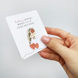 Puede incluir: Una tarjeta blanca con el texto "Los petirrojos aparecen cuando los seres queridos están cerca" y un pin de petirrojo dorado con corazones rojos.