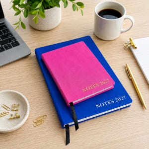 Puede incluir: Un cuaderno rosa brillante sobre un cuaderno azul más grande, ambos con el texto NOTES 2027 grabado en dorado. Cada cuaderno incluye un marcapáginas de cinta negra. Los cuadernos están sobre un escritorio con un portátil, una taza y clips.