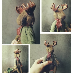 May include: A handmade moose toy with brown fur, a green sweater, and a red and white scarf. The moose is sitting on a wooden surface with green leaves.