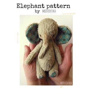 Op de afbeelding: Een pluche, beige olifantenspeelgoed met een lichtblauwe en bruine gepatenteerde stof op de oren. Het speelgoed wordt in de handen van een persoon gehouden.