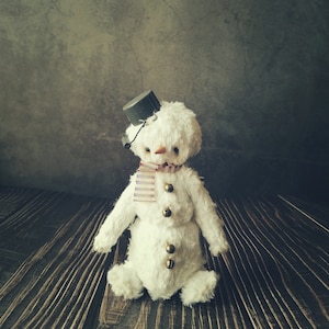 May include: A white plush snowman with a black top hat, a red and white striped scarf, and gold jingle bells. The snowman is sitting on a wooden surface.