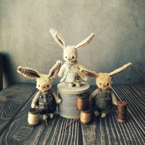 Puede incluir: Tres conejos de peluche con ojos marrones y orejas largas, que llevan monos y una camisa blanca, de pie sobre una superficie de madera con carretes de hilo.