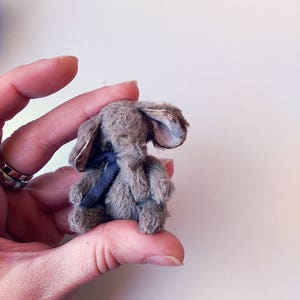 Puede incluir: Un pequeño elefante de peluche gris con una cinta azul alrededor del cuello. El elefante está sentado con las orejas levantadas.