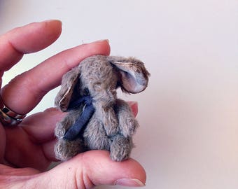 May include: A small gray plush elephant toy with a blue ribbon around its neck. The elephant is sitting up with its ears perked up.