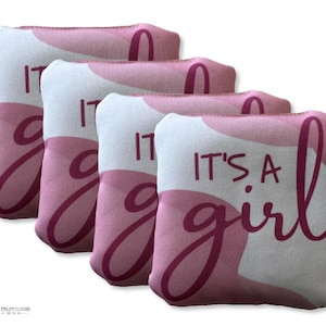 May include: Four pink and white bean bags with the text "IT'S A girl" in a cursive font. The bean bags have a pink and white abstract design. The bean bags are for a baby shower or gender reveal party.