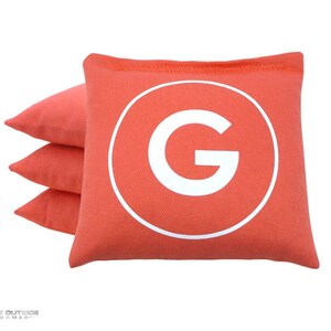 May include: Four coral-colored bean bags, three stacked and one in front. The front bean bag has a white circle with a large white "G" in the center. The bean bags are likely for a game like cornhole.