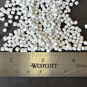 May include: Close-up of a pile of small, white, cylindrical plastic pellets. A stainless steel ruler with black markings and the "WESTCOTT" logo is in the foreground, measuring the pellets. The pellets are scattered on a dark surface.