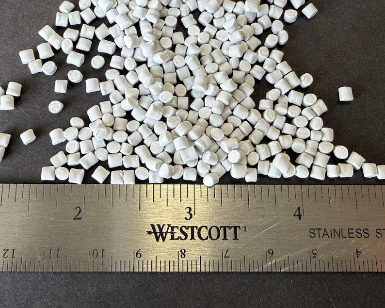 May include: Close-up of a pile of small, white, cylindrical plastic pellets. A stainless steel ruler with black markings and the "WESTCOTT" logo is in the foreground, measuring the pellets. The pellets are scattered on a dark surface.