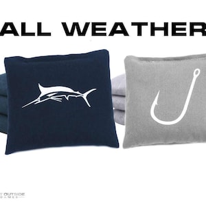 May include: Navy blue and gray cornhole bags with white graphics of a marlin and a fish hook. The text "ALL WEATHER" is displayed in black above the bags. The bags are designed for outdoor games.
