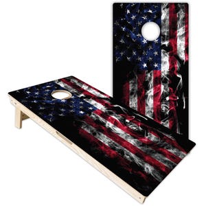 Könnte beinhalten: Ein Paar rechteckige Cornhole-Spielbretter aus Holz mit einem grafischen Druck der amerikanischen Flagge in einer dunklen, rauchigen Optik. Jedes Brett hat ein kreisförmiges Loch oben, helle Holzrahmen an den Seiten und ein klappbares Stützbein.