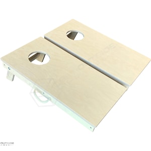 May include: Two wooden cornhole boards with a natural wood finish and white trim. Each board has a circular hole near the top. A white rope handle is attached to the bottom of one board. The boards are angled slightly, suggesting they are ready for play.
