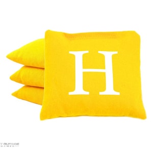 May include: A stack of four yellow bean bags. One bag features a large white letter "H". The bags are made of a textured fabric and are set against a white background. The bottom left corner has the text "GET OUTSIDE GAMES".