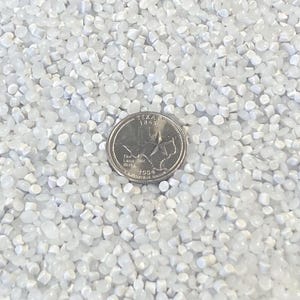 May include: A close-up shot of a pile of small, translucent, white plastic pellets. A U.S. quarter coin is in the center of the pellets for size comparison. The pellets are cylindrical and evenly distributed.
