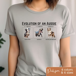 May include: A gray t-shirt with the text "Evolution of an Aussie" and three images of an Australian Shepherd dog at different ages. The text under the images reads "Aussie Baby", "Ausshhole", and "Australian Shepherd".