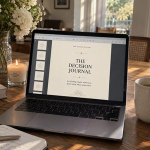 May include: A silver laptop displaying a digital document titled The Decision Journal by The Studio Within, featuring a cream-colored background with black serif text and a sunburst graphic at the bottom, centered on a wooden surface.