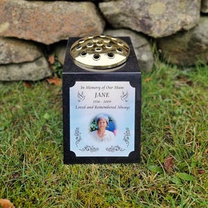 May include: Black granite memorial vase with a gold metal flower holder. The vase has a white plaque with the text "In Memory of Our Mum JANE 1936 - 2009 Loved and Remembered Always" and a photo of a woman.