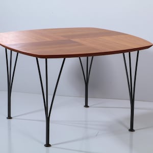 May include: A square wooden table with rounded corners and a warm brown wood grain finish. The table is supported by four black metal legs, each featuring a unique tripod-like structure that branches out from a single base point to the tabletop.