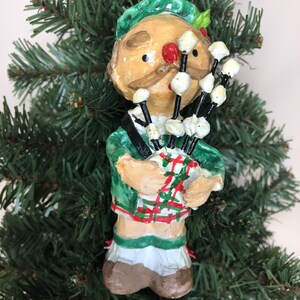 Peut inclure: Une d&eacute;coration de No&euml;l artisanale repr&eacute;sentant un homme portant un kilt vert et jouant des cornemuses. L'ornement est fait de papier m&acirc;ch&eacute; et a un style fantaisiste et caricatural.