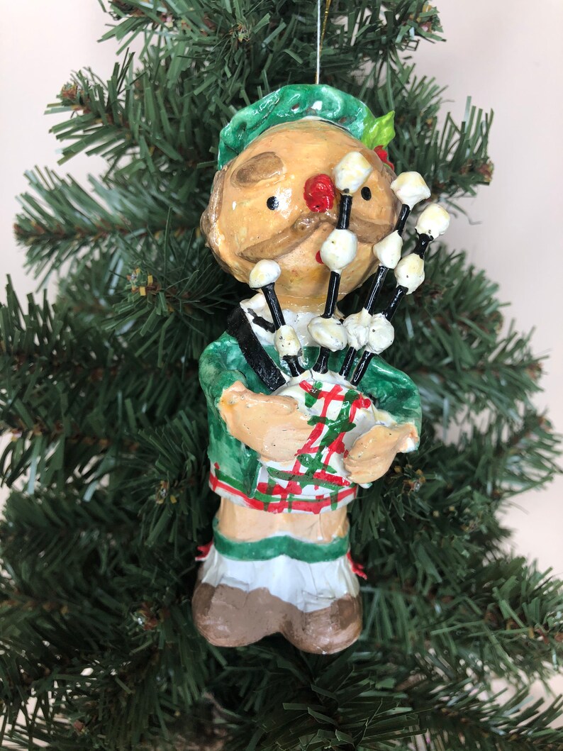 Peut inclure: Une d&eacute;coration de No&euml;l artisanale repr&eacute;sentant un homme portant un kilt vert et jouant des cornemuses. L'ornement est fait de papier m&acirc;ch&eacute; et a un style fantaisiste et caricatural.