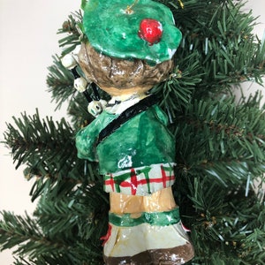 Peut inclure: Une d&eacute;coration de No&euml;l artisanale repr&eacute;sentant une personne portant un kilt vert et un chapeau vert avec un pompon rouge. La personne joue de la cornemuse.