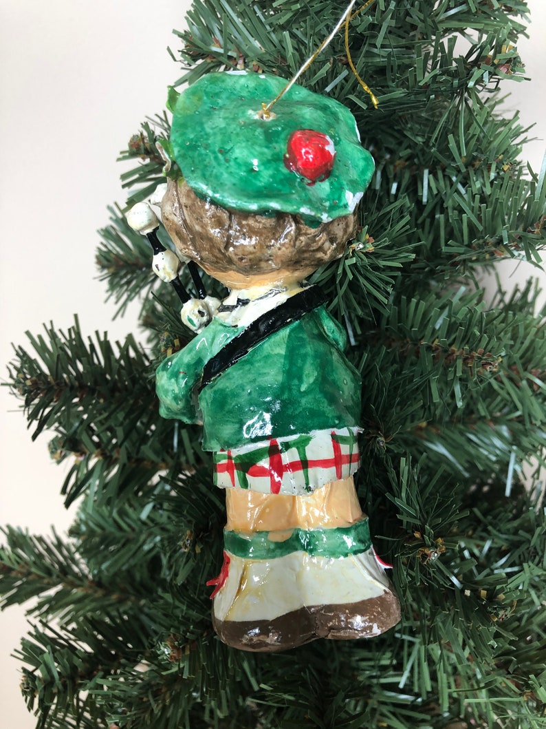 Peut inclure: Une d&eacute;coration de No&euml;l artisanale repr&eacute;sentant une personne portant un kilt vert et un chapeau vert avec un pompon rouge. La personne joue de la cornemuse.