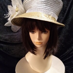 May include: A silver and gold hat with a large white flower and a veil. The hat is sitting on a white stand with a sparkly base.