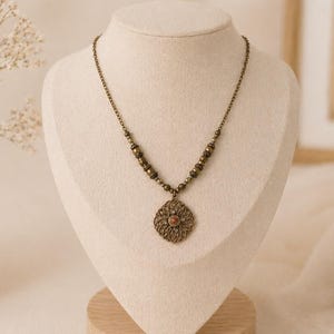 Könnte beinhalten: Halskette im Vintage-Stil mit einer bronzefarbenen Kette und Anhänger. Der Anhänger hat ein offenes Blumenmuster mit einem kleinen, rotbraunen Stein in der Mitte. Die Halskette wird auf einer cremefarbenen Büste präsentiert.