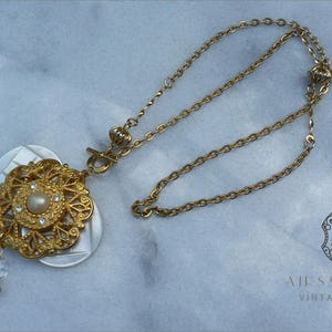 Peut inclure: Un collier en or avec un grand pendentif orné d'une perle et d'une coquille blanche. Le pendentif est attaché à une chaîne en or.